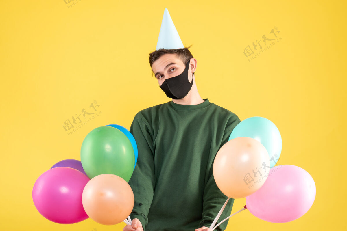 气球正面图：戴着派对帽和彩色气球的年轻人站在黄色的背景复制空间上帽子快乐年轻人