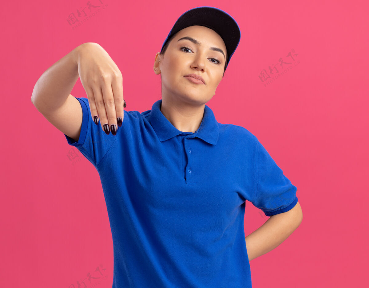 交货身穿蓝色制服 戴着帽子的年轻送货员自信地站在粉红色的墙上做手势年轻女人帽子