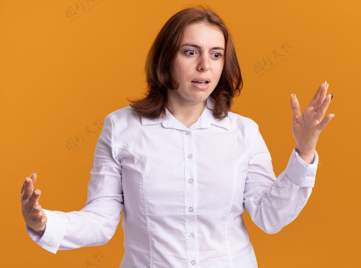 站着穿白衬衫的年轻女子一边看一边困惑地打着手势 双手站在橙色的墙上年轻手靠边
