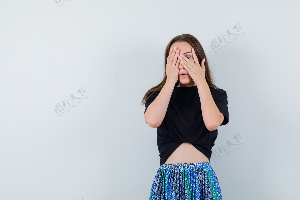 女性穿着黑色t恤和蓝色裙子的年轻女子用手捂住脸的一部分 透过手指看 看起来很迷人护理皮肤水疗