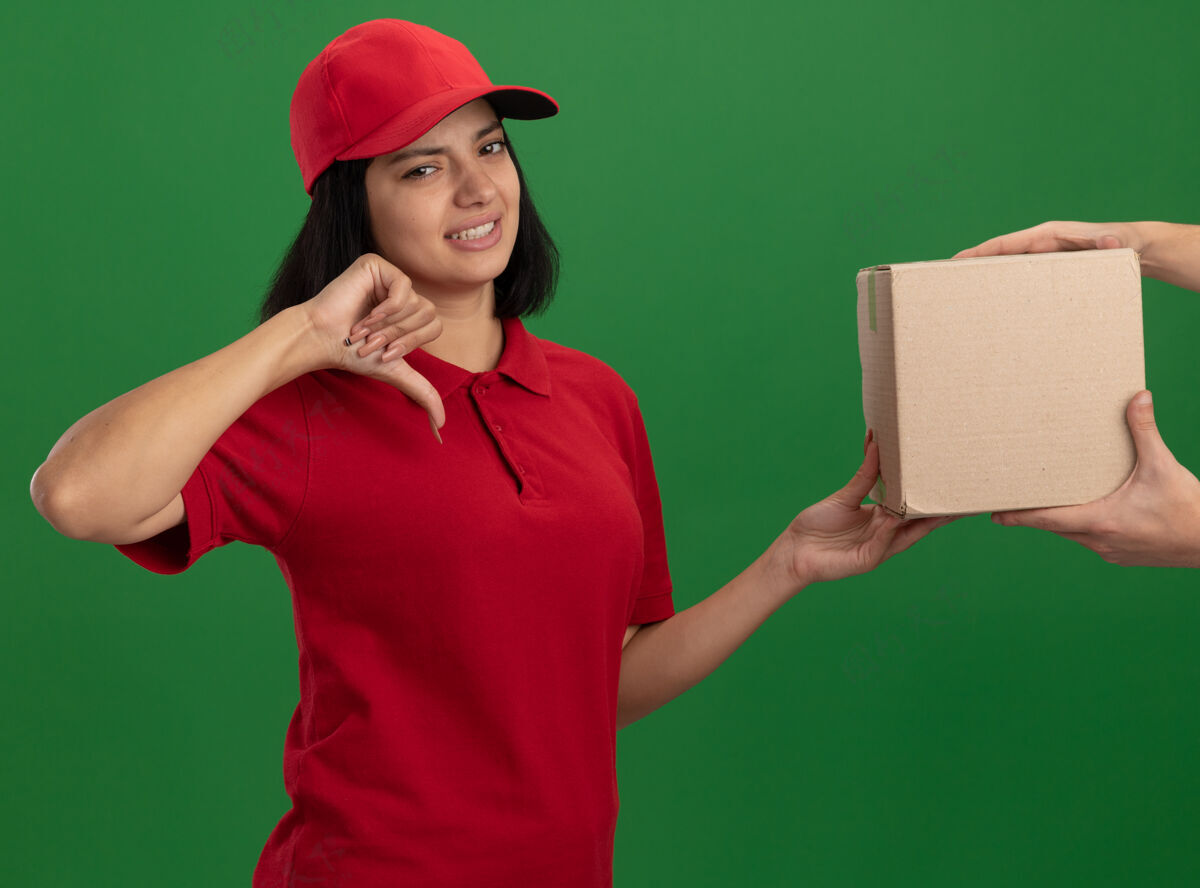 女孩身穿红色制服 头戴鸭舌帽的年轻送货员拿着纸板箱 站在绿色的墙上 看着一个看起来很不高兴 大拇指朝下的顾客放下帽子顾客