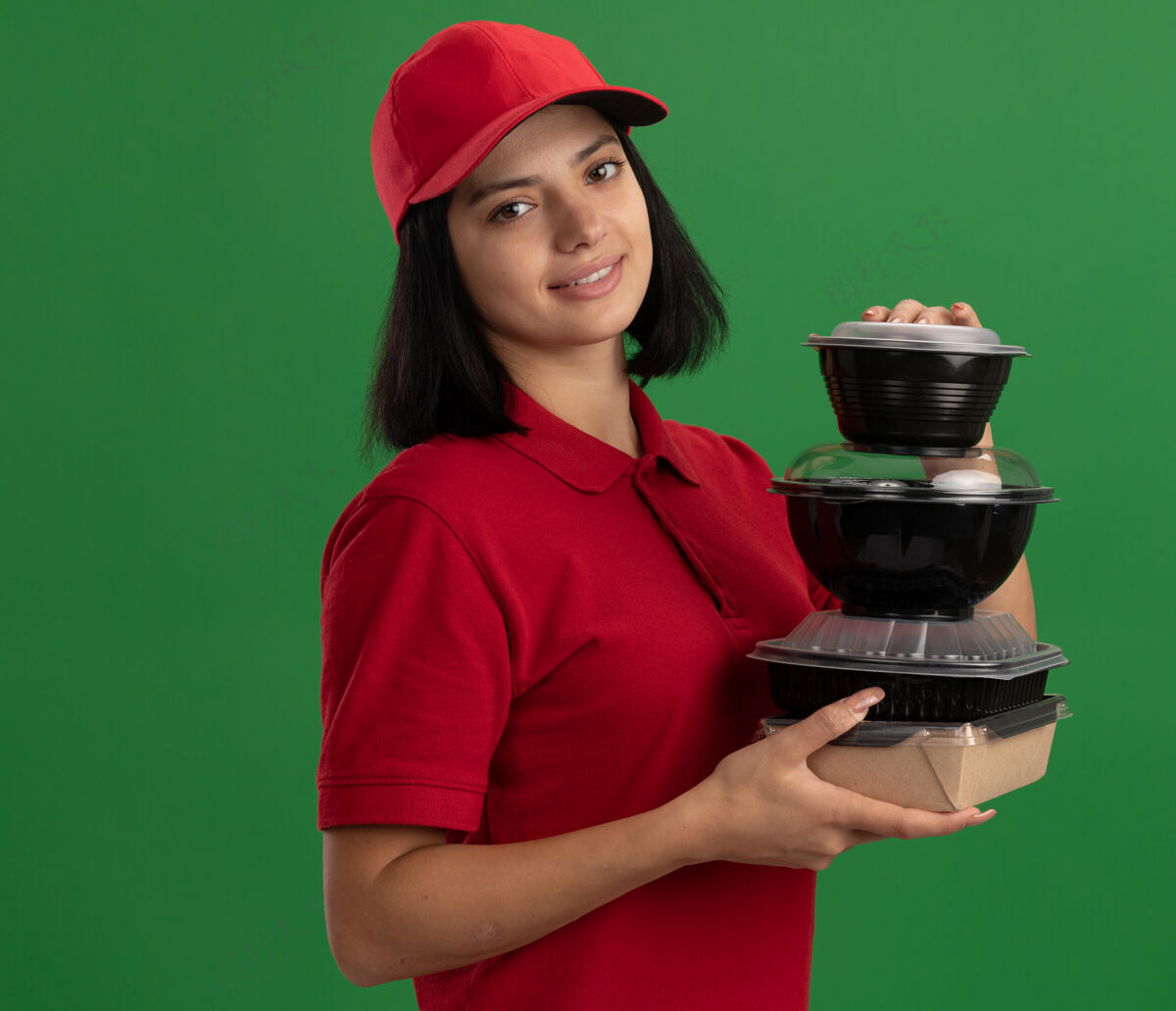制服穿着红色制服 戴着帽子 手里拿着一叠食品包装 微笑着友好地站在绿色墙壁上的快乐的年轻送货女孩拿着微笑年轻