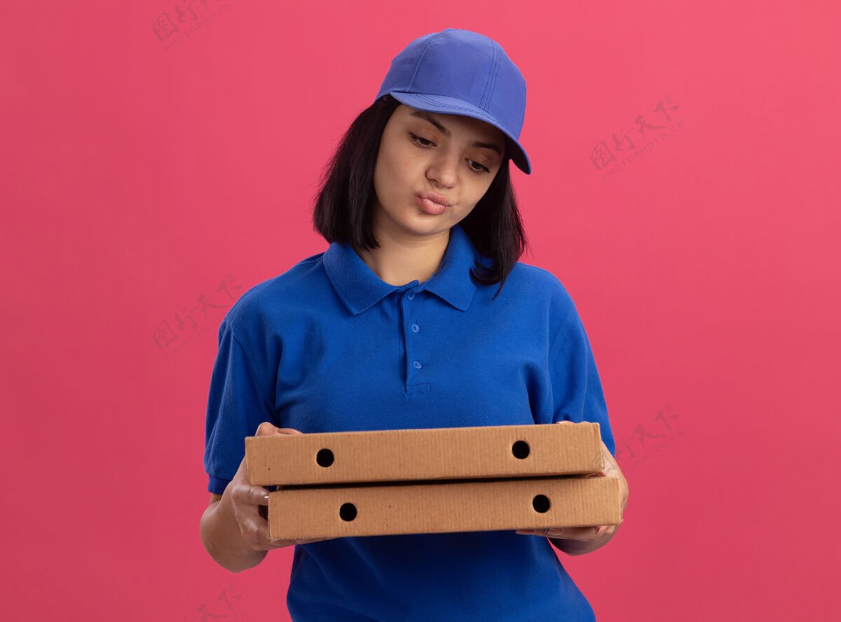站着身穿蓝色制服 头戴鸭舌帽 拿着披萨盒的年轻送货员站在粉色的墙上 神情不悦 表情悲伤女孩制服悲伤