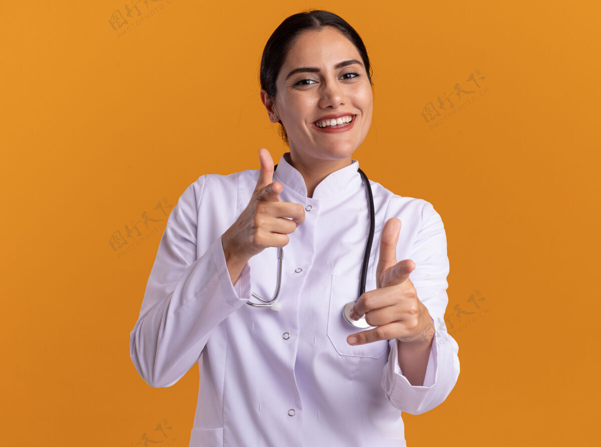 指快乐的年轻女医生 穿着医用外套 脖子上戴着听诊器 面带微笑 食指指着前面站在橙色的墙上外套医学年轻