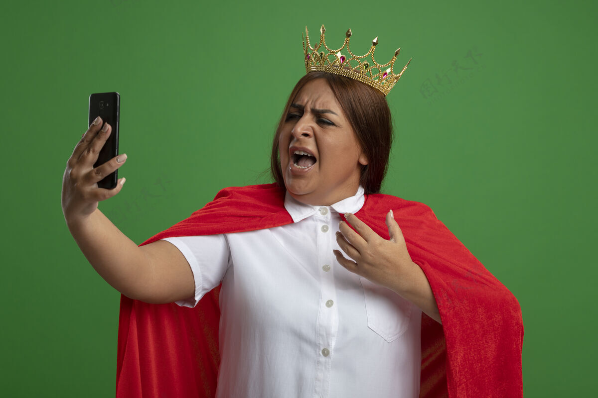 女性愤怒的中年女超人戴着王冠 手持手机看着绿色背景上的孤立超级英雄电话皇冠