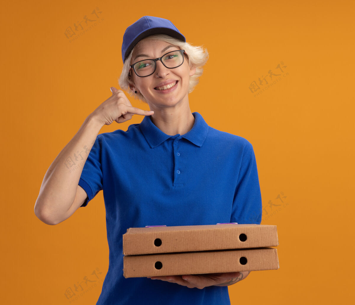 女人身着蓝色制服 戴着眼镜 拿着披萨盒 微笑着做着“我微笑”手势的年轻送货员 站在橙色的墙上穿制服帽子