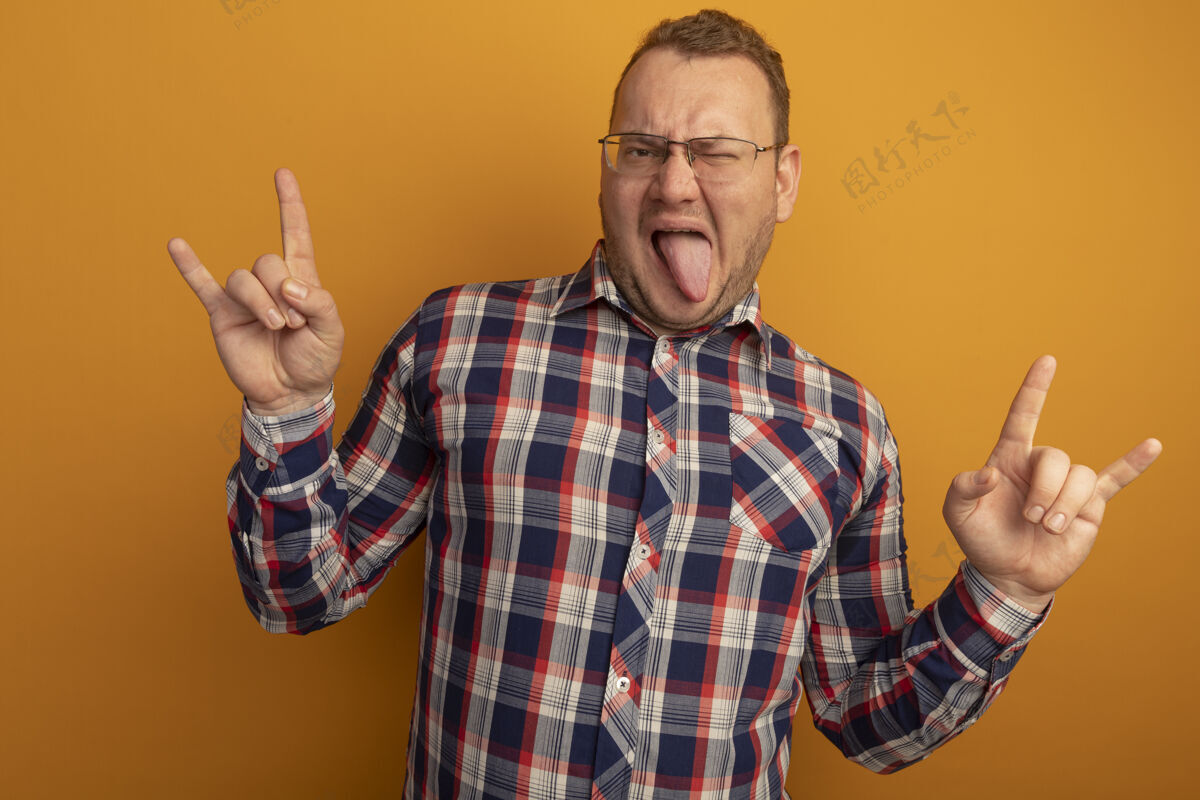 舌头戴着眼镜 穿着格子衬衫的快乐男人快乐快乐地展示着站在橙色墙壁上伸出舌头的岩石符号男人坚持欢呼