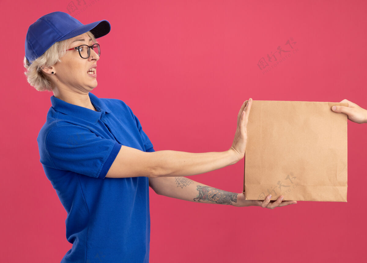 站着身穿蓝色制服 戴着帽子的年轻送货员站在粉色的墙上拒绝拿纸包裹年轻女人制服