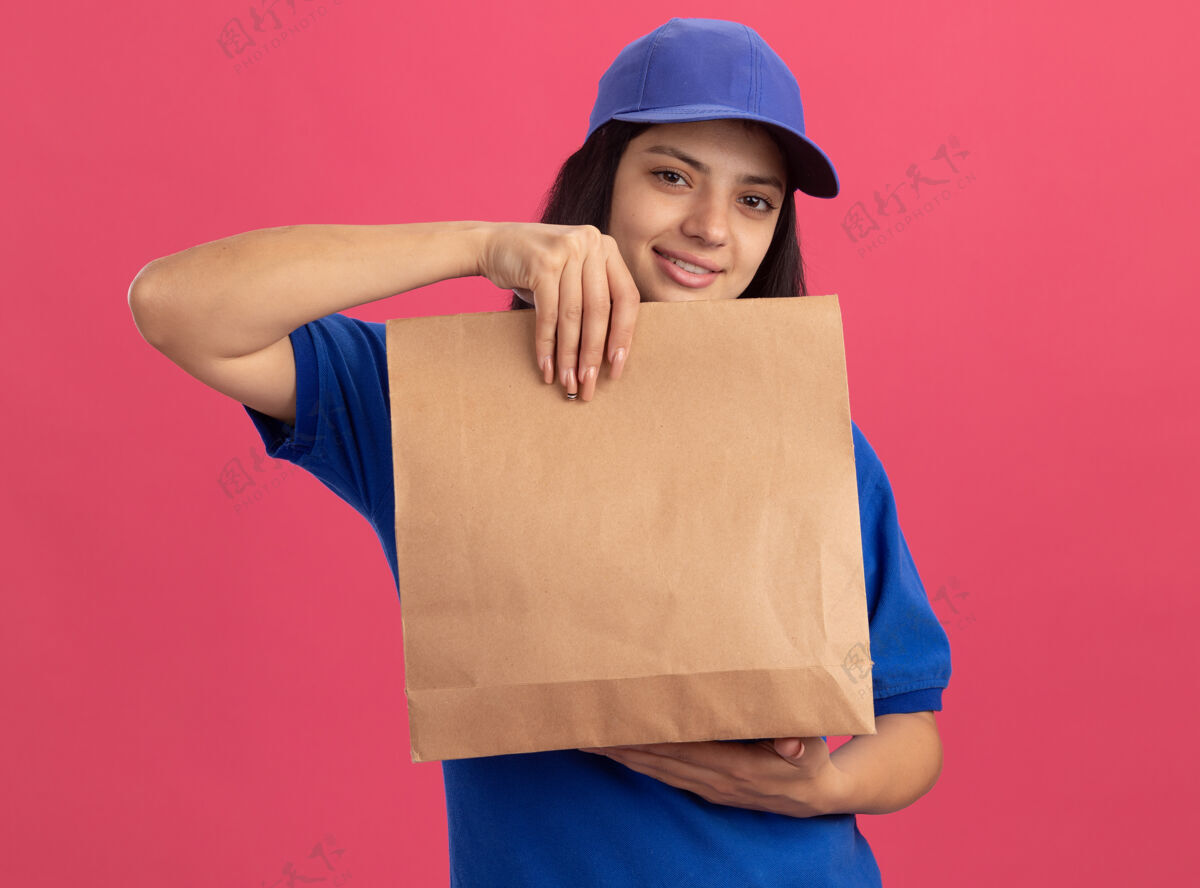 包装身穿蓝色制服 头戴礼帽的年轻送货女孩站在粉红色的墙上 露出自信的微笑年轻送货女孩