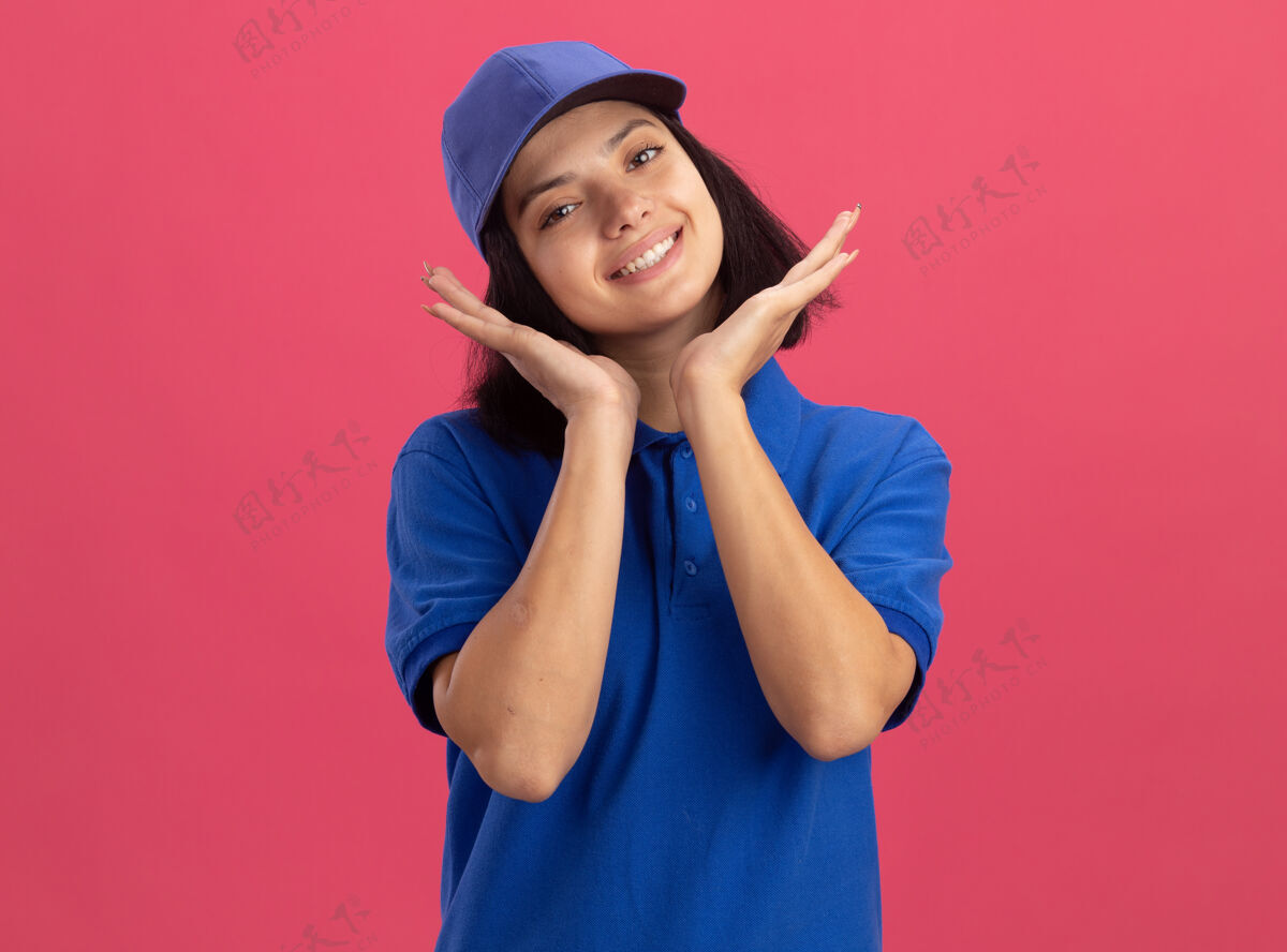 年轻身着蓝色制服 头戴鸭舌帽 站在粉色墙壁上微笑的快乐的年轻送货女孩站立制服递送