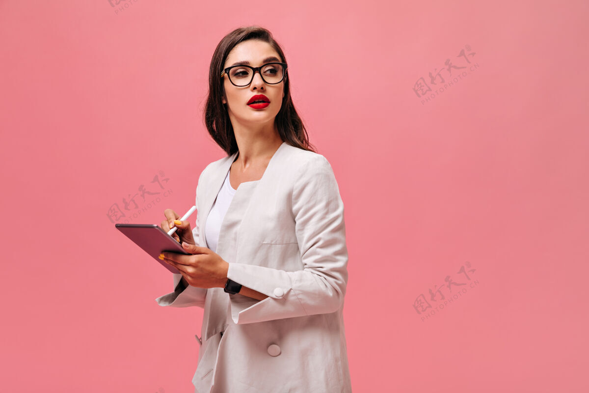 姿势戴眼镜的女人在平板电脑上做笔记漂亮的深棕色头发 大红唇 穿着浅色套装 在孤立的背景上摆姿势粉色休闲站立