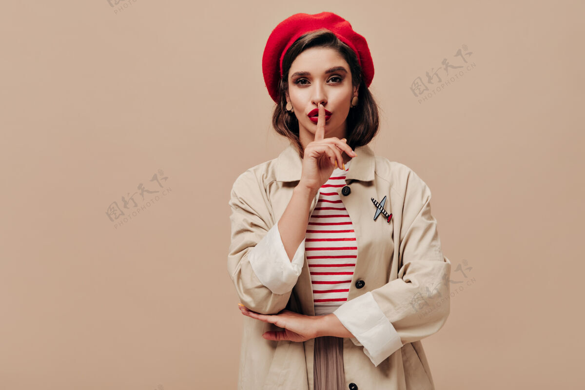 肖像身着红色贝雷帽和米色战壕的女士要求在与世隔绝的背景下保守秘密严肃的年轻女子 嘴唇明亮 戴着时髦的帽子摆姿势米色背景休闲姿势