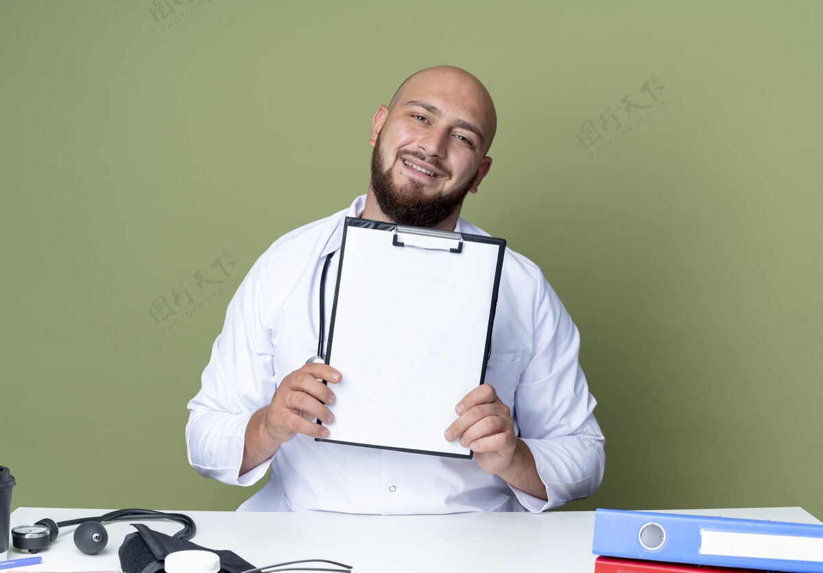 男性高兴的年轻秃头男医生穿着医用长袍和听诊器坐在办公桌前 手持医疗工具 把剪贴板隔离在绿色背景上请坐着工具