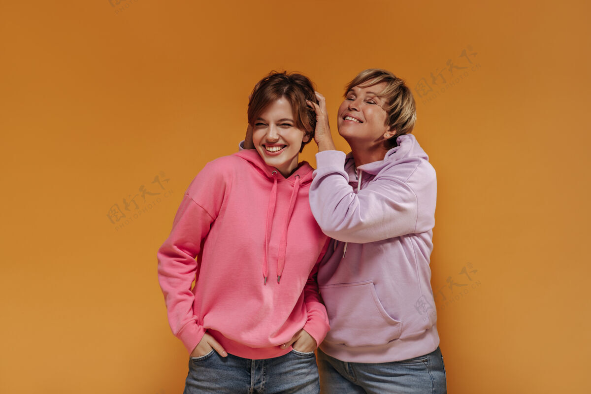 年轻欢快的两位女士 短发 宽大时尚的连帽衫 酷酷的牛仔裤 在橙色孤立的背景下微笑着 玩得很开心老年人肖像室内