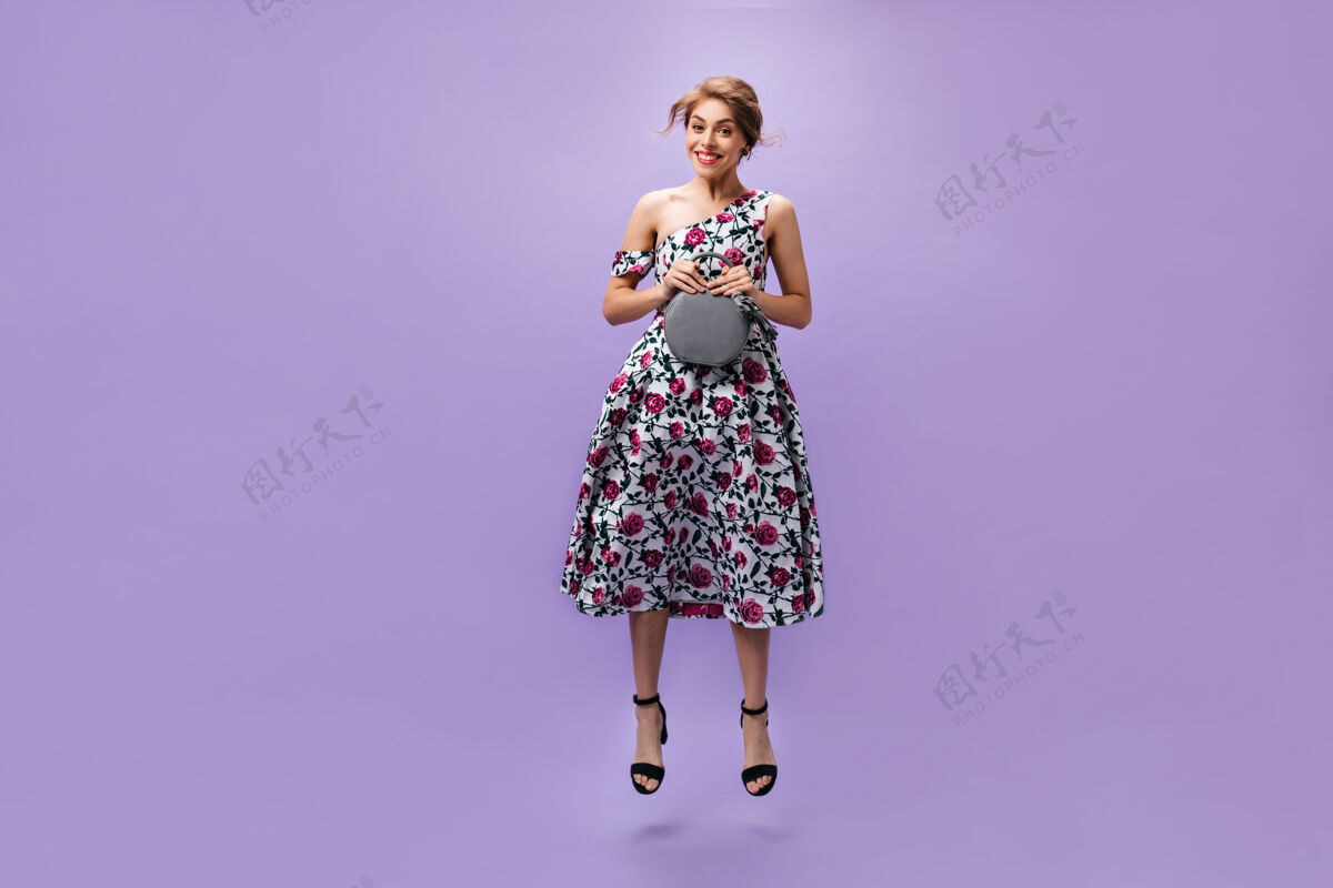 女性身着迷笛裙的微笑女士拿着手提包 在紫色的背景上跳跃穿着凉服的漂亮年轻女子拿着灰色的包摆姿势紫色女人脸