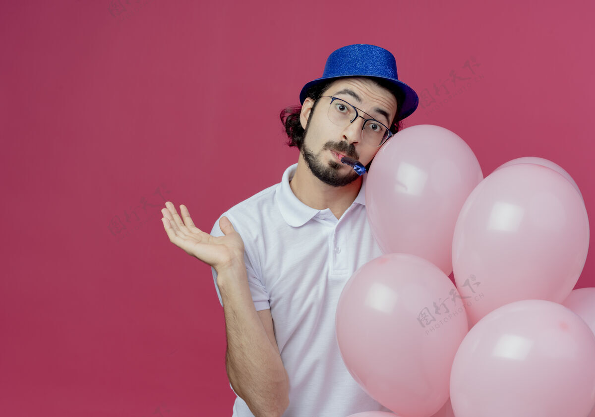 气球高兴的帅哥戴着眼镜 戴着蓝色帽子 手持气球吹着口哨 摊开手 孤立地在粉色背景上复制空间手穿手持