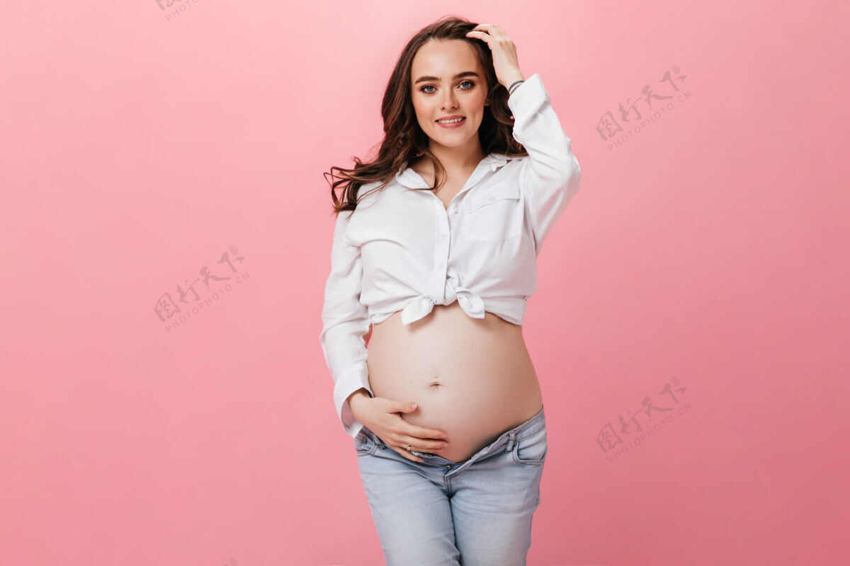 怀孕身穿白色短款衬衫和牛仔裤的黑发孕妇在孤立的粉色背景下抚摸着腹部微笑腹部为人父母父母