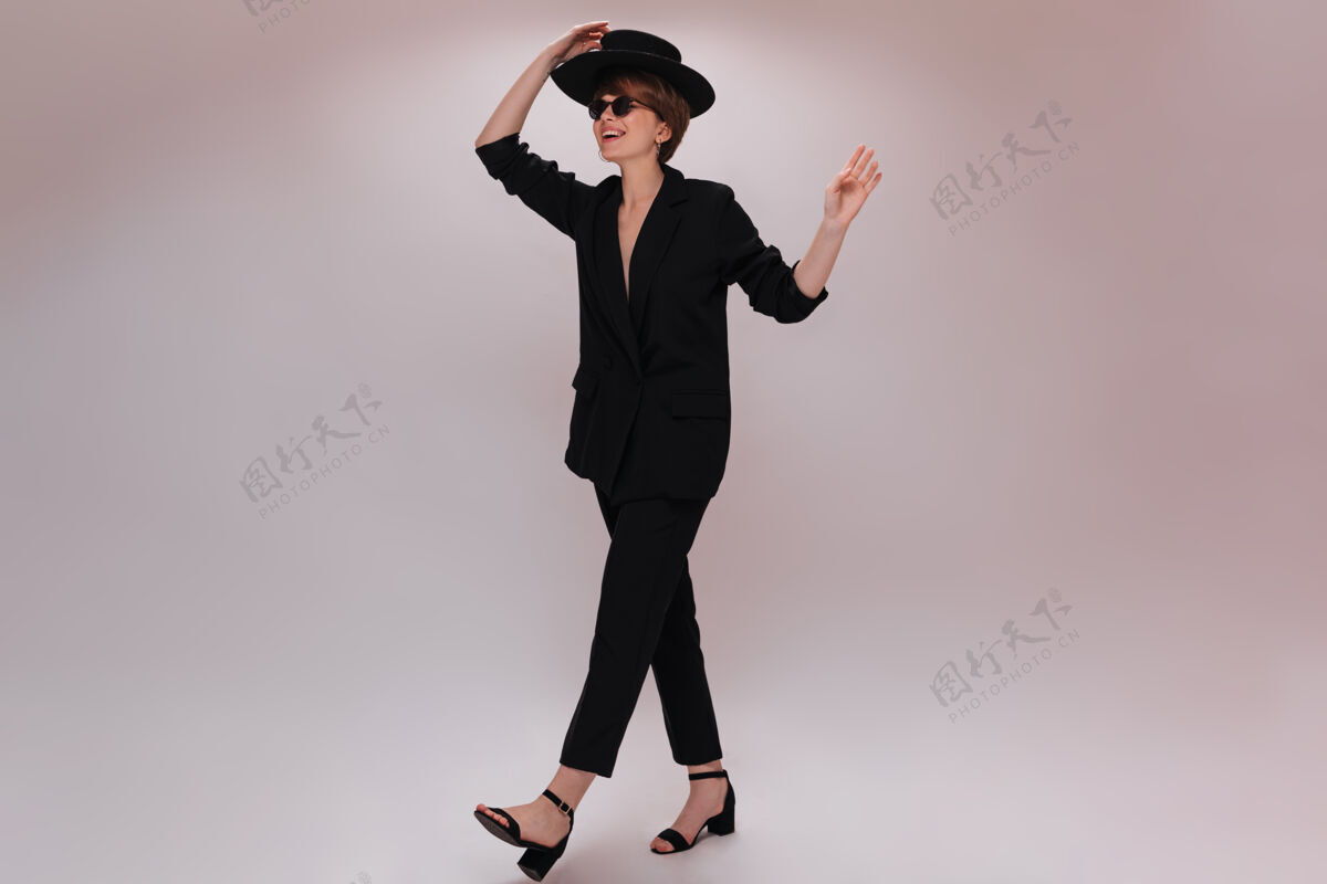 女性穿着西装戴着帽子的年轻女子在孤立的背景上移动穿着黑色夹克和裤子的迷人短发女士在白色背景上行走年轻女孩短发