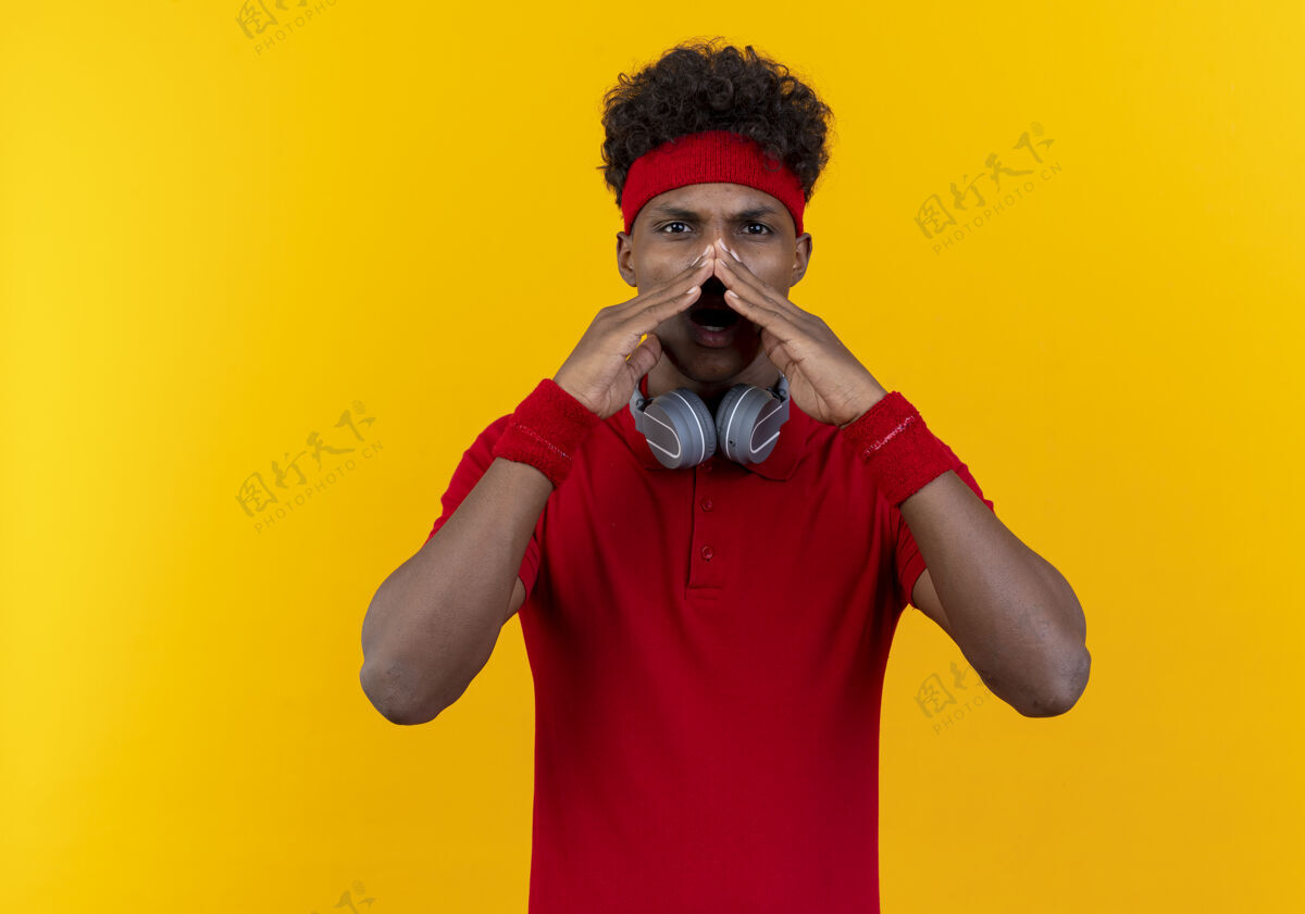 运动看着镜头 年轻的美国黑人运动男子戴着头带和腕带 脖子上戴着耳机 在黄色背景下呼唤着一个孤立的人非裔美国人穿相机