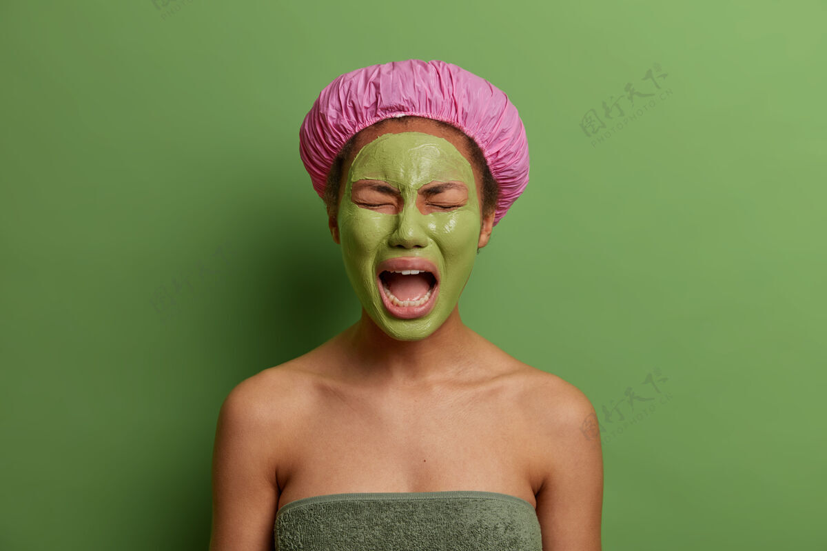 美容师哭哭啼啼的女人厌倦了在spa沙龙的美容 张大嘴巴 戴着完美皮肤的绿色面膜 戴着浴帽和毛巾围着身体 靠着墙站着浴缸肖像美容