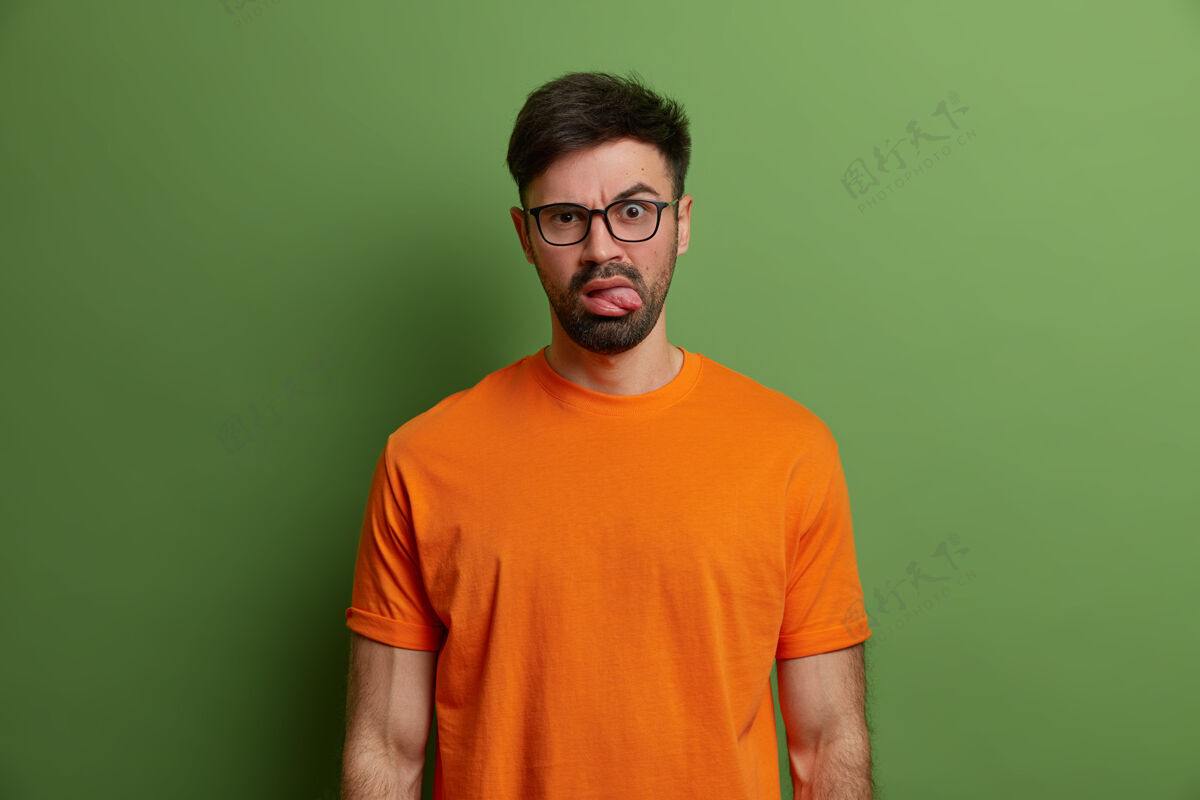 男人不满未刮胡子的欧洲年轻人伸出舌头 做出烦躁不安的表情 戴着透明眼镜和亮橙色t恤 隔着亮绿色的墙深色表演眼镜