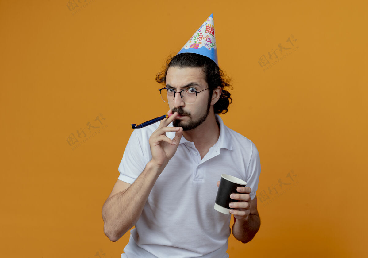 帽子愤怒的帅哥戴着生日帽吹着口哨 手里拿着一杯孤立在橙色背景上的咖啡生气杯子拿着