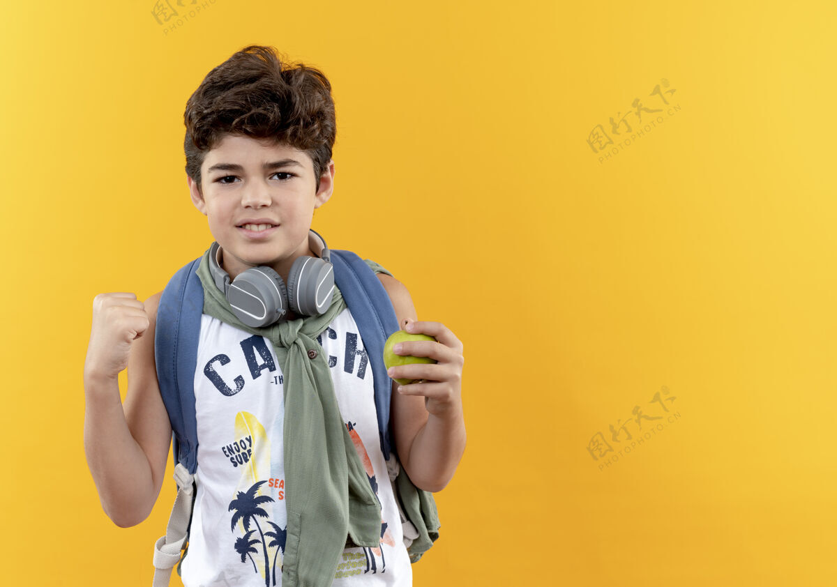 小高兴的小男孩戴着书包和耳机拿着苹果和显示是的手势隔离在黄色背景与复制空间展示拿着背