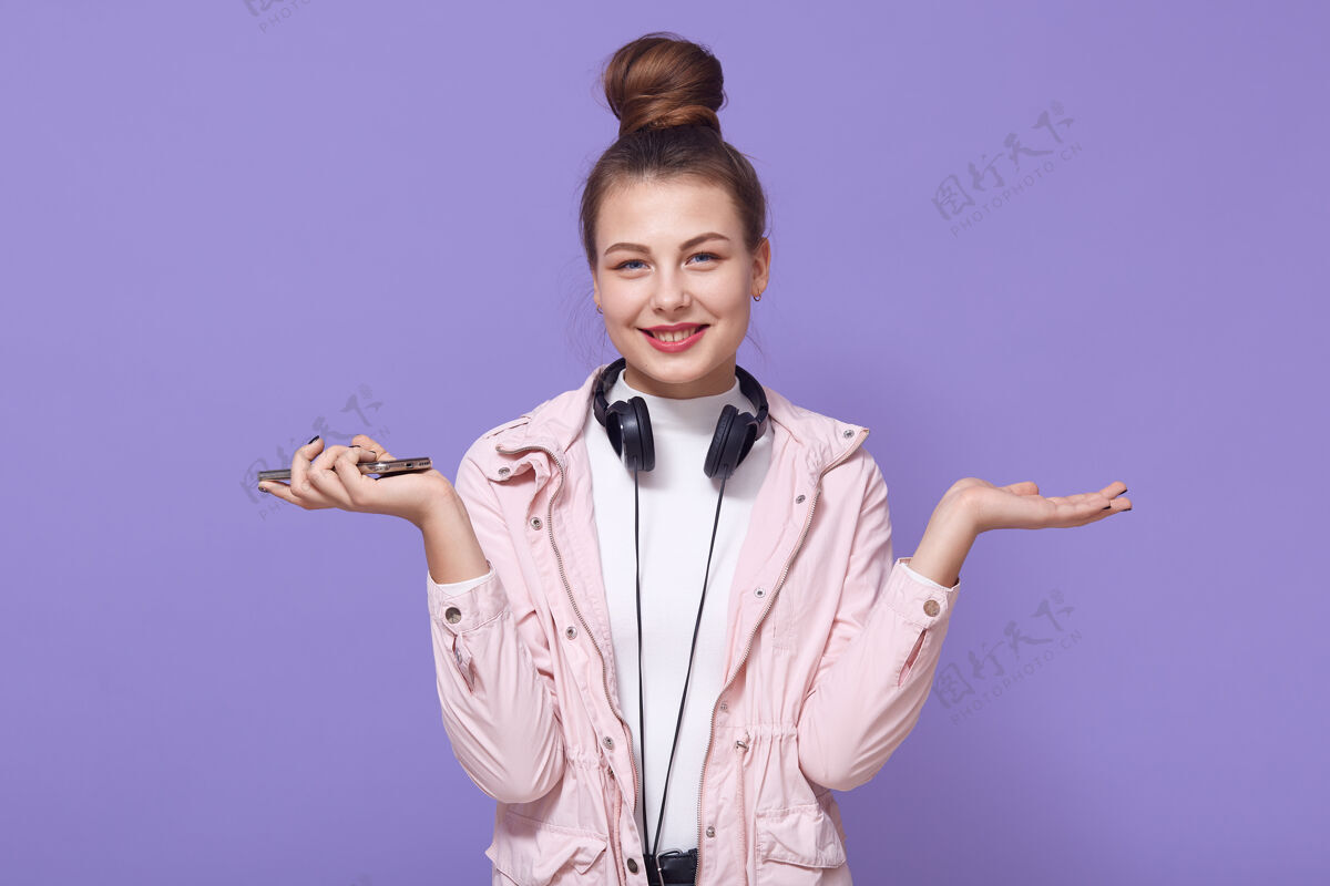 姿势穿着浅粉色夹克的迷人的欧洲年轻女孩站在淡紫色的墙上 脖子上戴着耳机 双手摊开 手机放在一边 带着迷人的微笑室内夹克女人