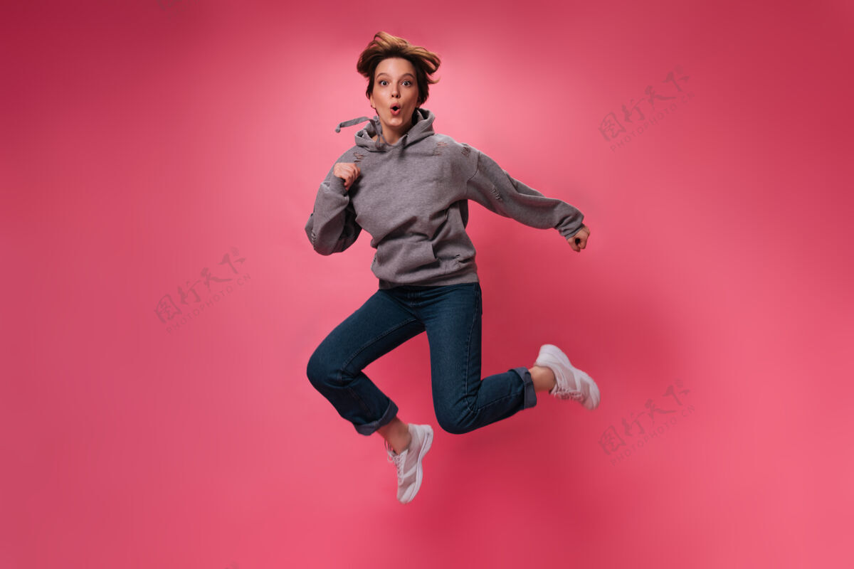 女性活泼的女人穿着舒适的衣服在粉色背景上跳跃迷人的女孩穿着连帽衫和牛仔裤的肖像在孤立地移动享受移动牛仔裤