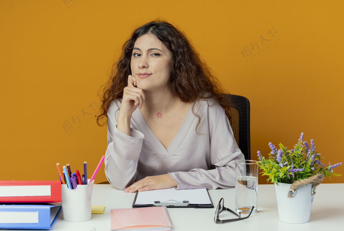 办公室自信的年轻漂亮的女上班族坐在办公桌前 手放在橙色背景的下巴上女性桌子年轻