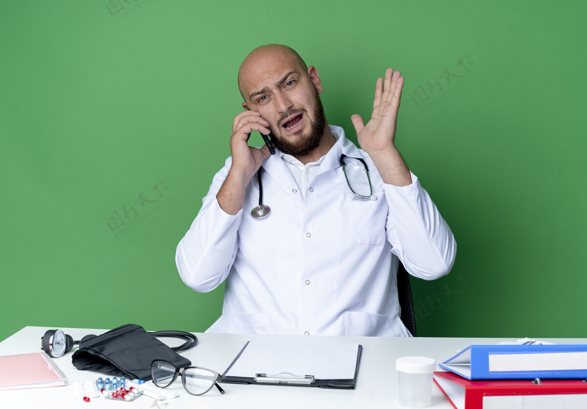 说话愤怒的年轻男医生穿着医用长袍 手持听诊器坐在办公桌旁 拿着医疗工具讲着电话 在绿色背景上摊开隔离的手听诊器坐着桌子