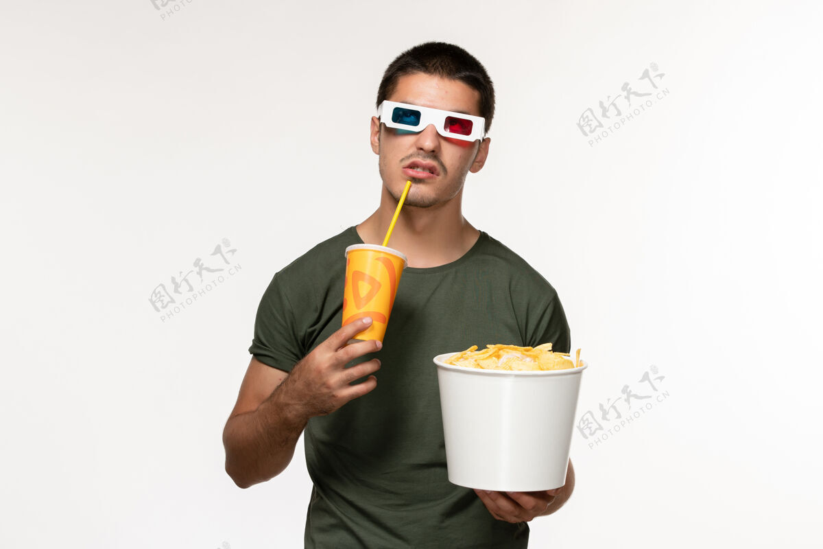 土豆正面图身穿绿色t恤的年轻男性手持土豆cips 戴着d太阳镜在白色墙壁上喝着苏打水电影男性孤独电影微笑年轻男性苏打水