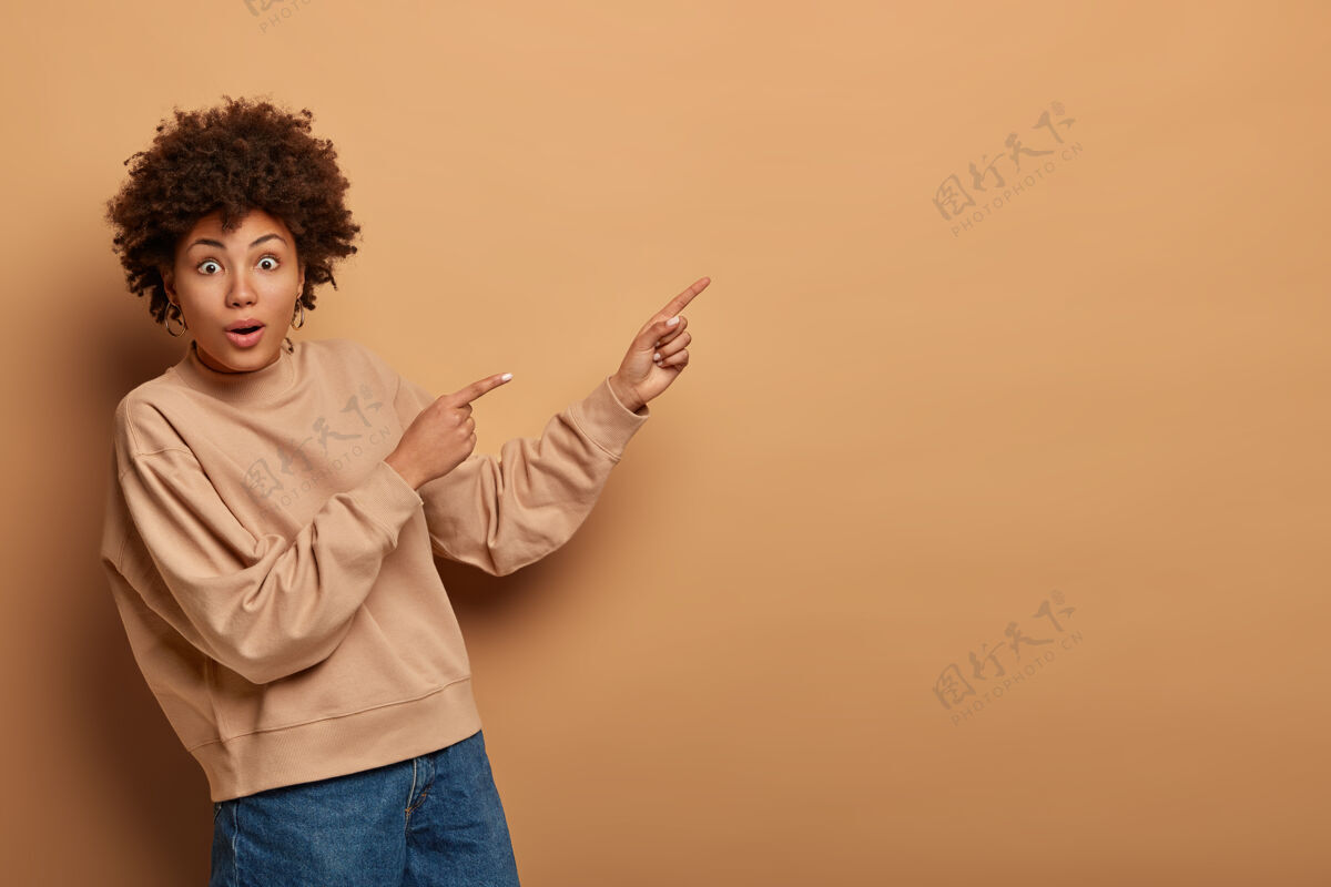 凝视好奇的卷发女人有好奇兴奋的目光 在空地上展示产品 给出建议 穿着棕色运动衫 说点击链接 孤立在米色墙上指向广告毛衣