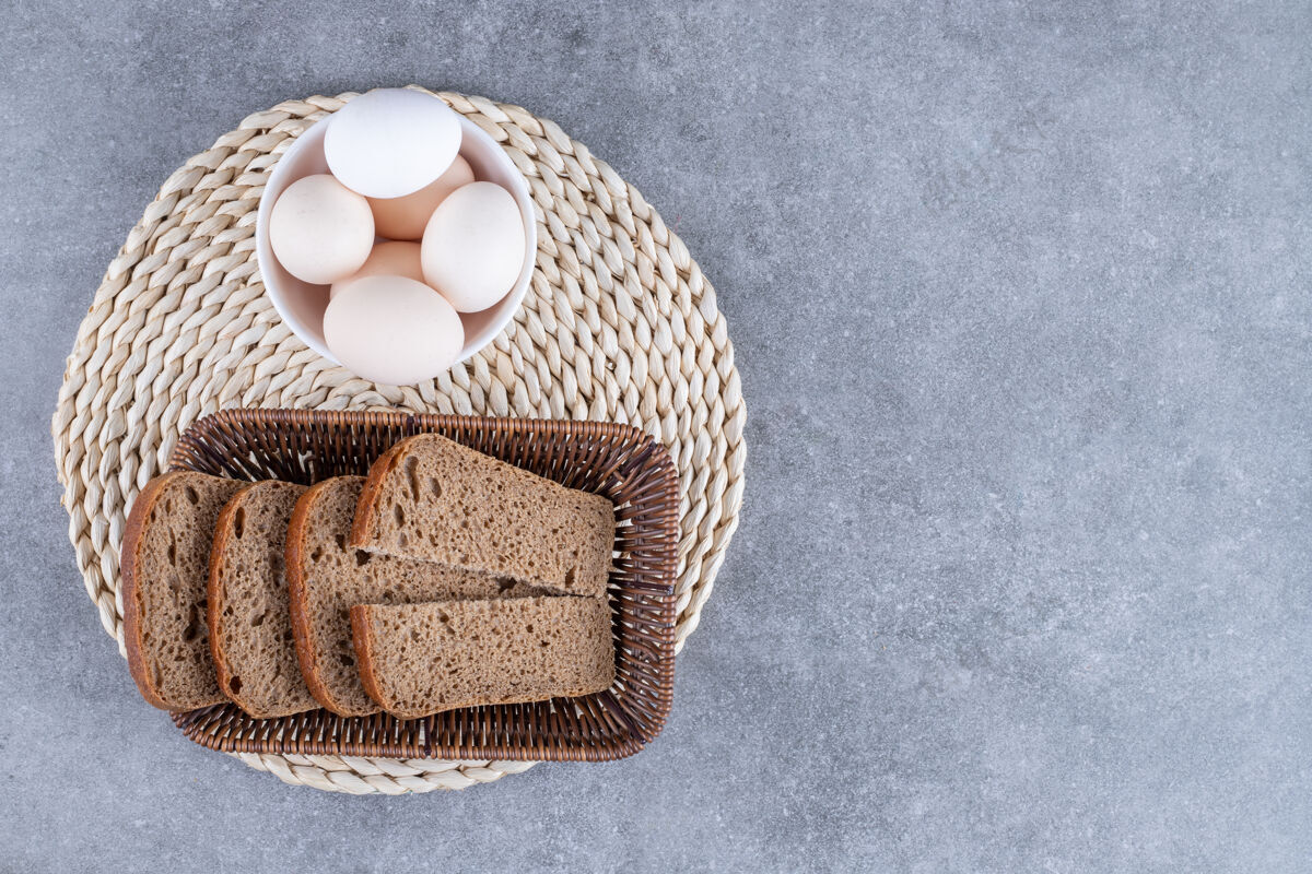 面包房柳条篮子的黑麦面包和一碗生鸡蛋放在石桌上糕点面包早餐