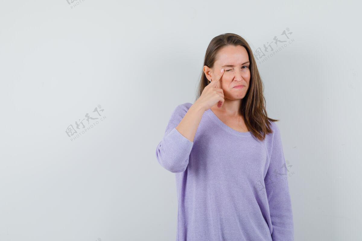 女士年轻的女性指着她的眼睑 一边眨着眼睛 一边穿着淡紫色的衬衫 看起来很专注优雅而卷发