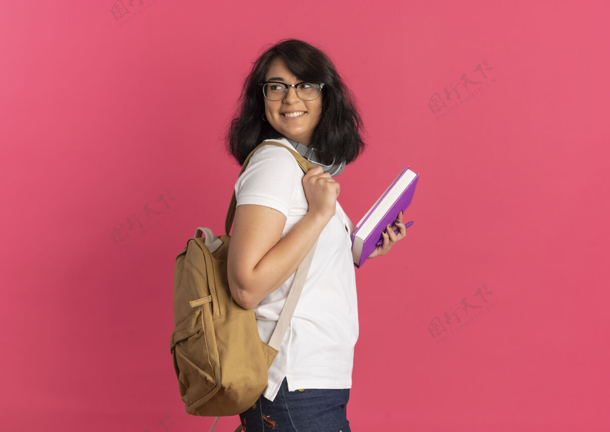 侧着年轻笑容可掬的白种女学生戴着眼镜 背着书包站在一旁 背着书包 拿着粉红色的书 留着复印空间漂亮包微笑