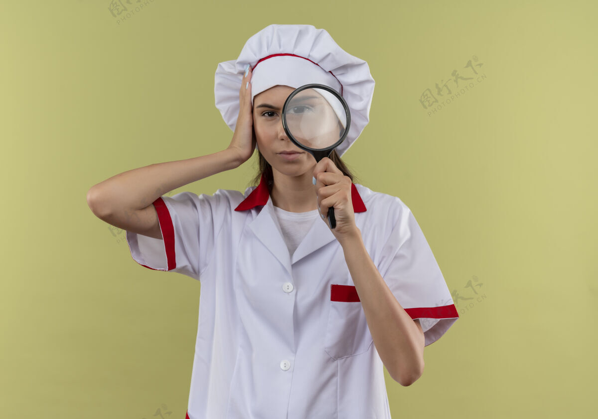 持有身着厨师制服的年轻自信的白人厨师女孩手持放大镜 将手放在有复印空间的绿地上手放大镜头