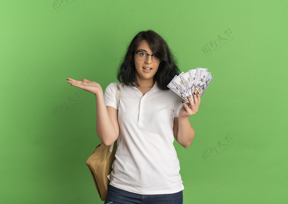 手年轻漂亮的白人女生戴着眼镜 背着书包 手里拿着钱 手伸直放在绿色的地方 留着复印空间拿眼镜绿色