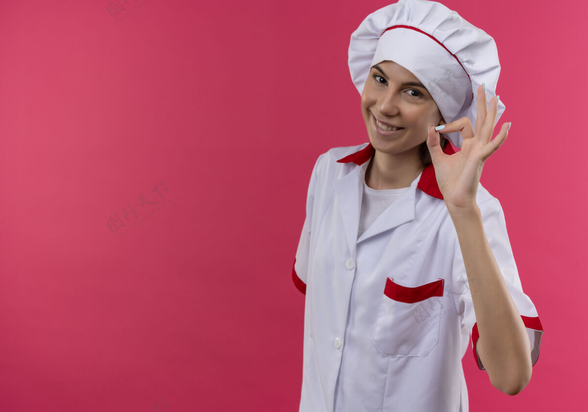 手势身着厨师制服 面带微笑的白人年轻厨师女孩摆出“ok”手势 看着粉色的相机 并留有复印空间粉红复制女孩
