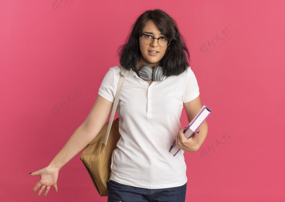 年轻年轻焦急漂亮的高加索女学生戴着眼镜 背着包 脖子上戴着耳机 手里拿着粉红色的书 还有复印空间女学生空间戴