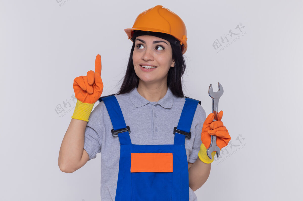 建筑工人身穿施工制服 头戴安全帽 戴橡胶手套 手持扳手 食指 微笑着站在白色墙壁上的年轻建筑工人安全显示橡胶