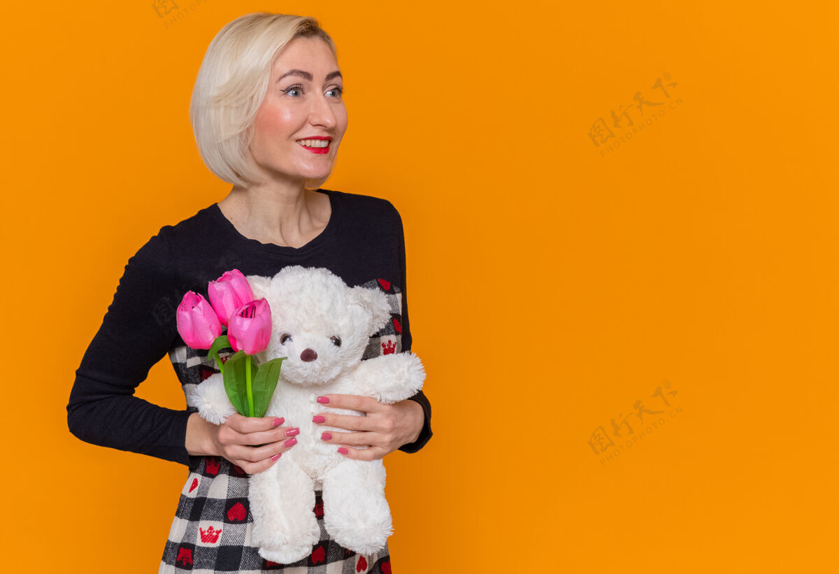 庆祝身着漂亮连衣裙的快乐年轻女子手持一束郁金香和泰迪熊作为礼物 站在橙色的墙上 微笑着欢庆国际妇女节年轻熊国际