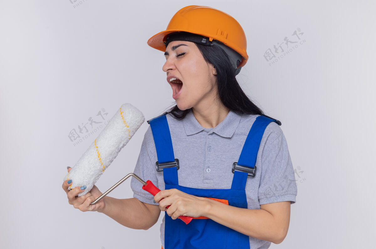 女人身穿施工制服 戴着安全帽 拿着油漆辊的年轻建筑工人 拿着油漆辊当麦克风 站在白色的墙壁上尽情歌唱麦克风头盔立场