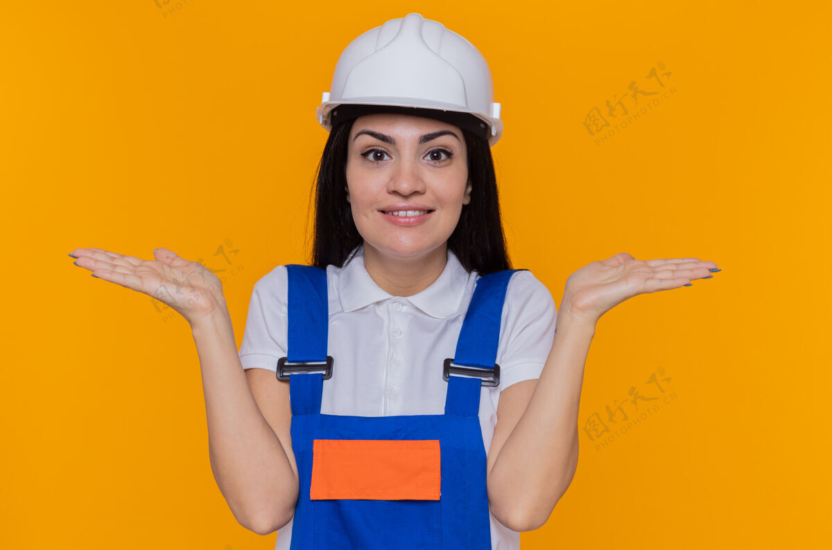 女人身穿施工制服 头戴安全帽的年轻建筑工人望着前方 面带微笑 兴高采烈地张开双臂站在橙色的墙上制服手臂微笑