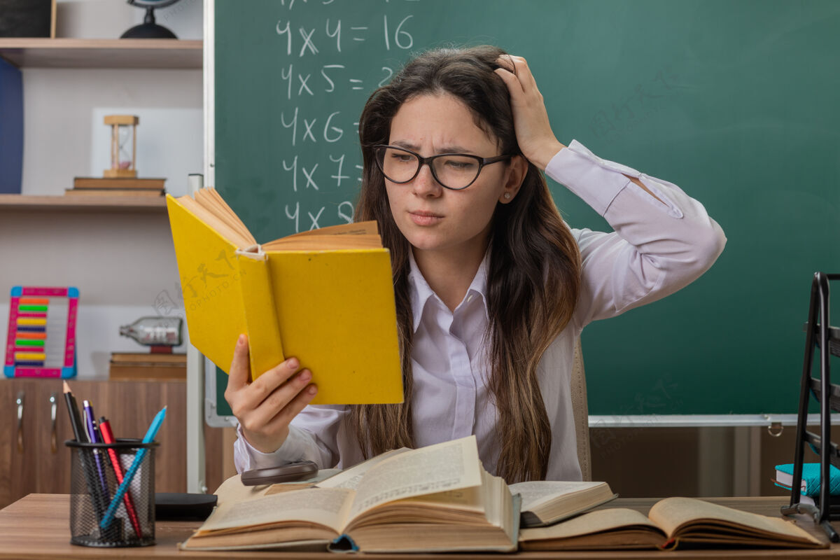 坐着戴着眼镜看书的年轻女老师正困惑地把手举在头上坐在教室黑板前的课桌旁阅读黑板年轻