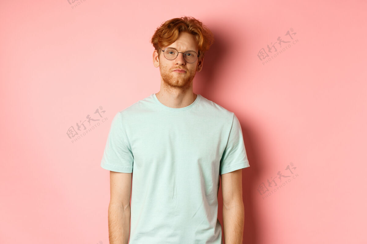 学生愁眉苦脸的红胡子男人 穿着t恤 戴着眼镜 站在粉色背景下 无聊而不习惯地盯着镜头休闲T恤红发