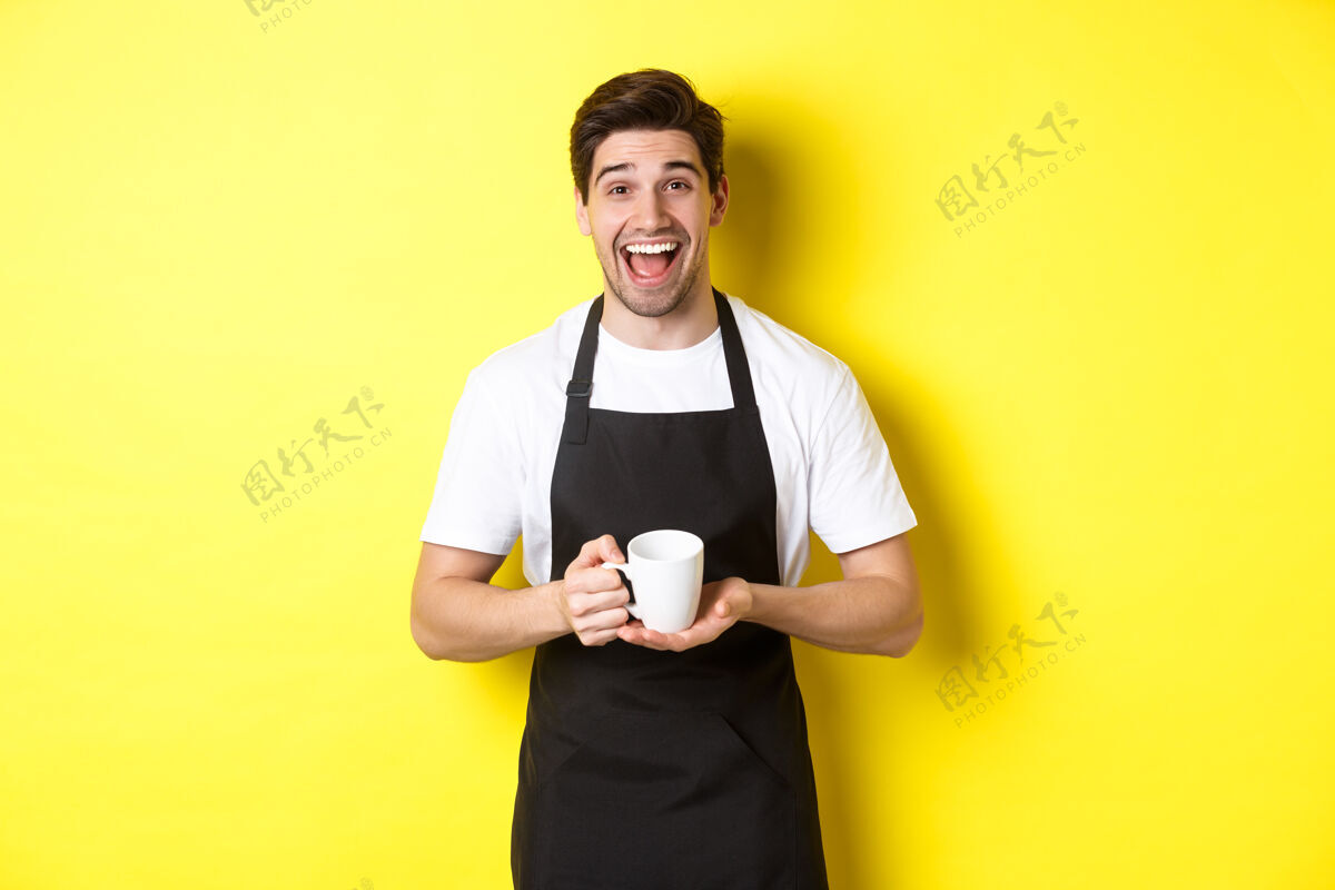 黄色快乐的咖啡师 穿着黑色围裙 手里拿着咖啡杯 站在黄色的背景上大笑男人男孩烹饪