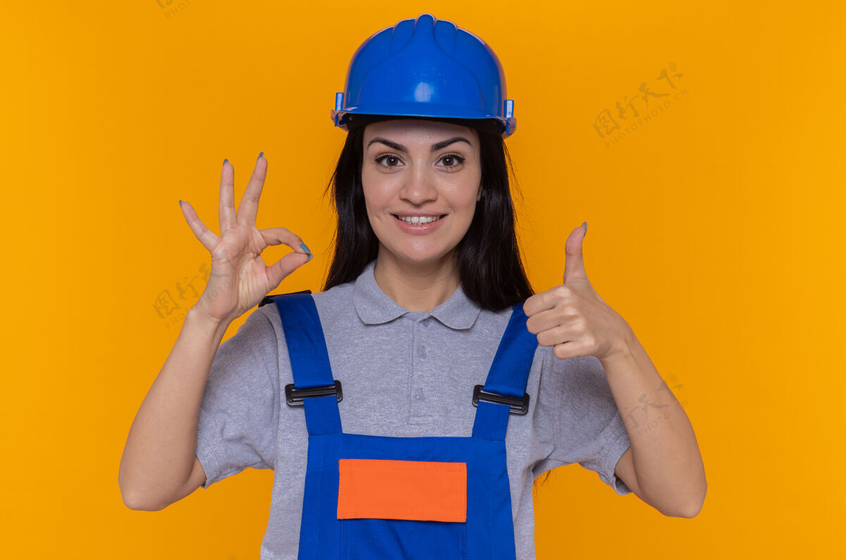 积极身穿施工制服 头戴安全帽的年轻建筑工人高兴而积极地向上竖起大拇指 橙色的墙上立着“ok”标志女人快乐年轻