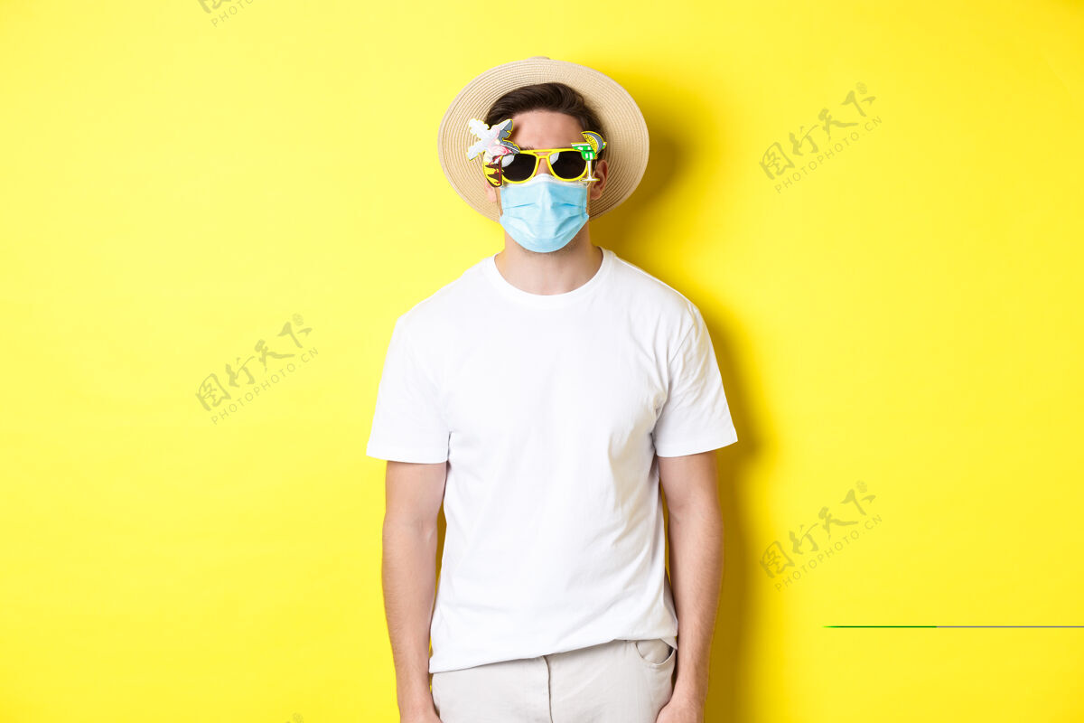 男人covid-19的概念 度假和社交距离戴着医疗面罩和带太阳镜的夏季帽子的男性游客 在大流行期间旅行 黄色背景面膜表情外科面膜