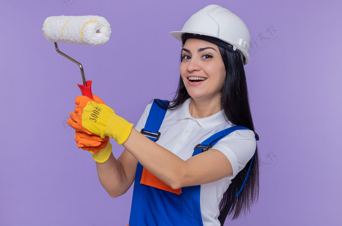 快乐身着施工制服 戴着安全帽 戴着橡胶手套 拿着油漆辊 兴高采烈地站在紫色墙壁上的快乐的建筑女青年穿上女人欢呼